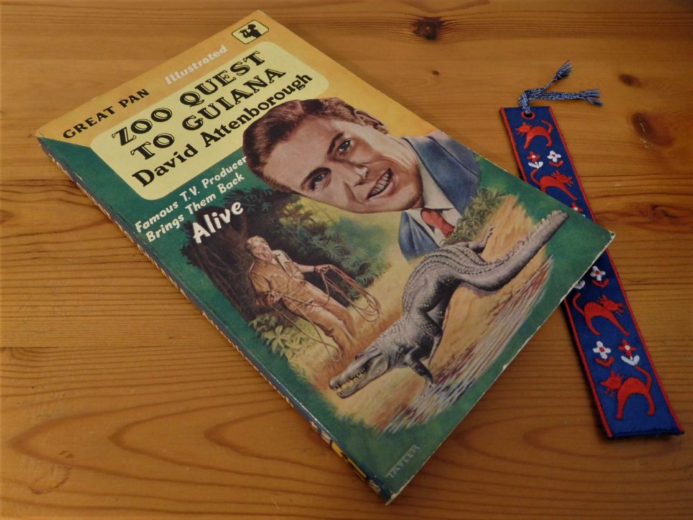 My paperback copy of 'Zoo Quest to Guiana' by David Attenborough (Pan, 1956), seen on a wooden table top next to a blue embroidered bookmark with red cats and red and white flowers. The green and yellow book cover is illustrated with two colour drawings of a young David Attenborough; one a head and shoulders portrait of him in a blue suit and red tie; the other of him in a beige safari suit, holding a lasso-like coil of rope, while approaching a crocodile beside a river with a lush jungle-like background. Posted today, 8 May 2025, on the occasion of Attenborough's 99th birthday.