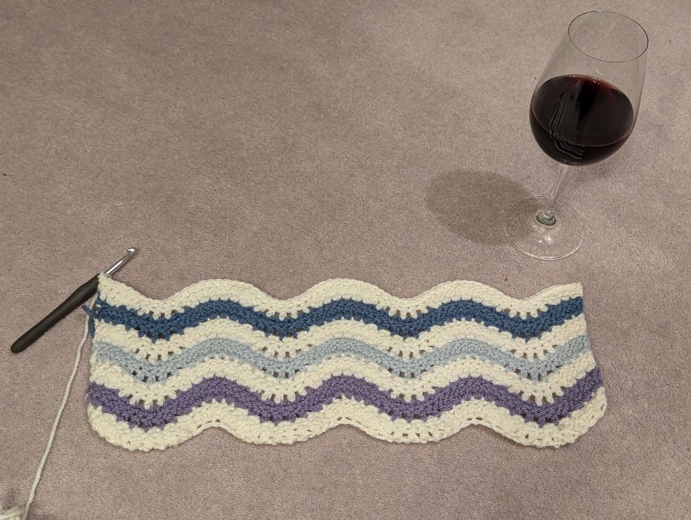 A half finished crochet blanket with the crochet hook still attached, made up of cream, purple, light blue and dark blue waves, sitting on the floor next to a glass of red wine. 