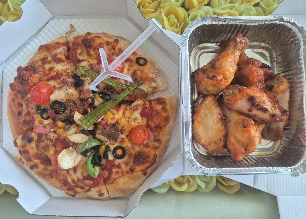 A 4-slice, personal-size "the works" pizza next t 6 buffalo wings/sticks inside a rectangular foil container