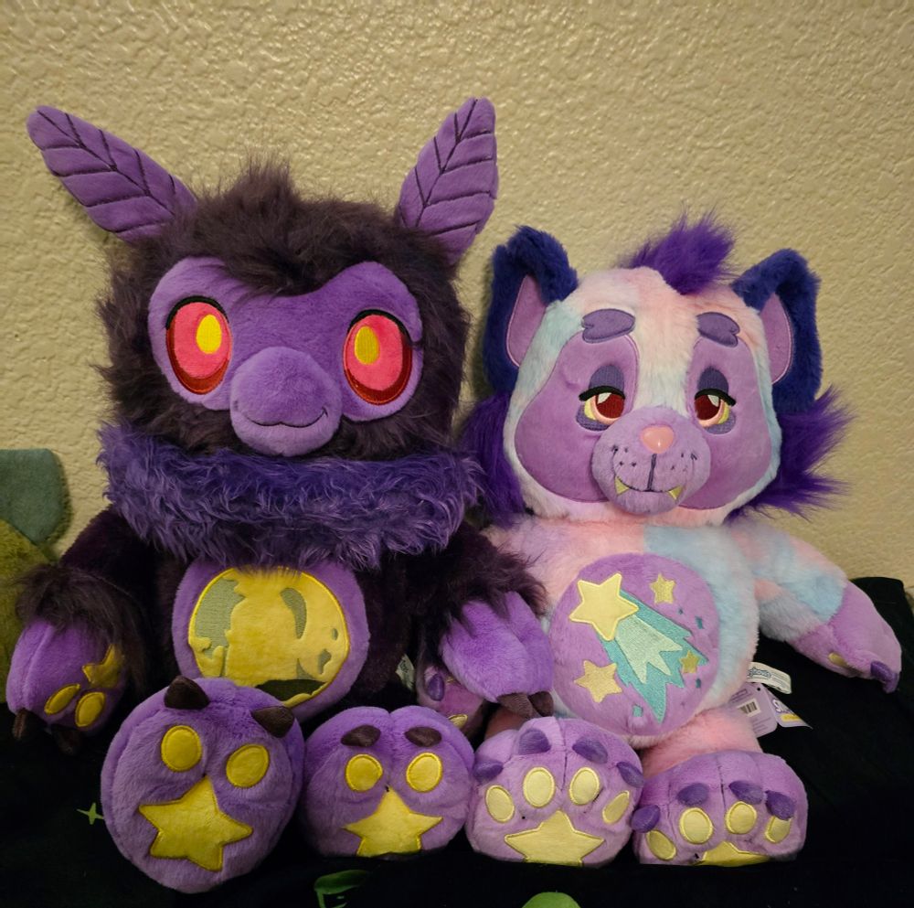Two plush toys are sitting side by side against a textured beige wall. They are detailed with fantasy and celestial themes.

The plush on the left resembles a dark purple moth creature with large, red eyes that have a yellow glow. It has a soft, fluffy mane and a rounded snout with a small smile. Its ears are long and pointed with a stitched pattern resembling moth antenna. The belly of the plush features an embroidered patch of a glowing yellow full moon with. Its paws have star and circle patterns embroidered in yellow on the purple fabric.

The plush on the right is a pastel-colored werewolf creature with a dreamy appearance. Its fur is a mix of soft pink, light blue, and lavender, with dark purple fluffy tufts on its cheeks and ears. It has a relaxed, half-lidded expression with red eyes and a tiny fang sticking out from its mouth. The belly features an embroidered design of a shooting star with a light blue trail. Like the moth plush, its paws also have embroidered star and circle patterns in yellow on purple fabric.

Both plush toys have unique celestial and whimsical details, with vibrant colors and embroidery enhancing their fantastical designs. They are placed on a black surface, contrasting with their colorful appearance.