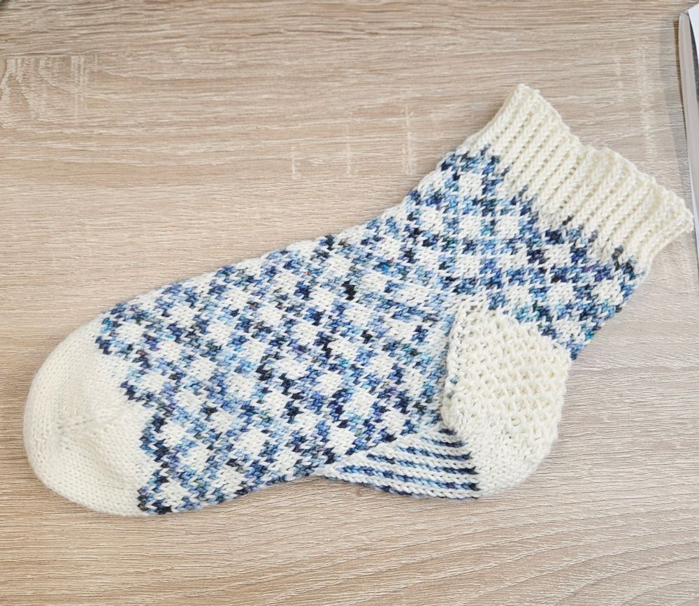 A completed colorwork sock done up in undyed white for the toes, background color, heel, and cuff, and a variegated blue. The colorwork is in a familiar gingham-esque diamond pattern. The pattern is Rhubarb and Custard by ZaneteKnits