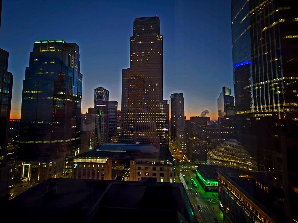 The Minneapolis city skyline at sunrise. Buildings appear dark with lights on in sporadic windows. The street is dotted wire light from cars and businesses. The sunrise on the horizon breaks through thr darkness in the background with an orange contrast. The sky is a deep shade of blue above the buildings. 