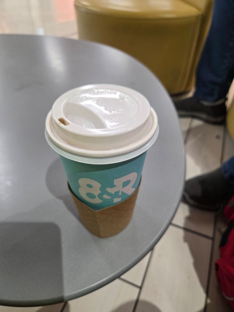 A to-go cup of coffee sits atop a grey table. The cup is light blue with a white lid and the cardboard coffee collar is down to show the shop logo. This cup is a vanilla oat milk latte from 8&R coffee shop in the Nashville Airport. 