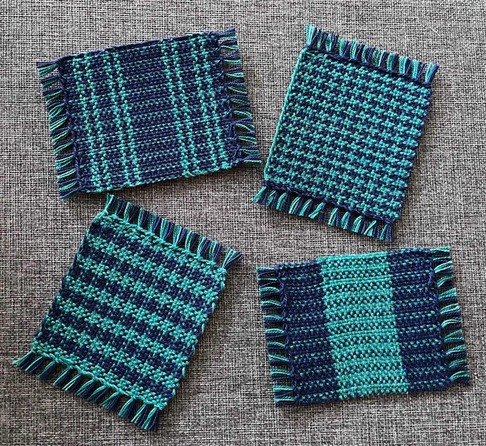 Four square woven coasters with fringe in shades of dark blue and turquoise.
