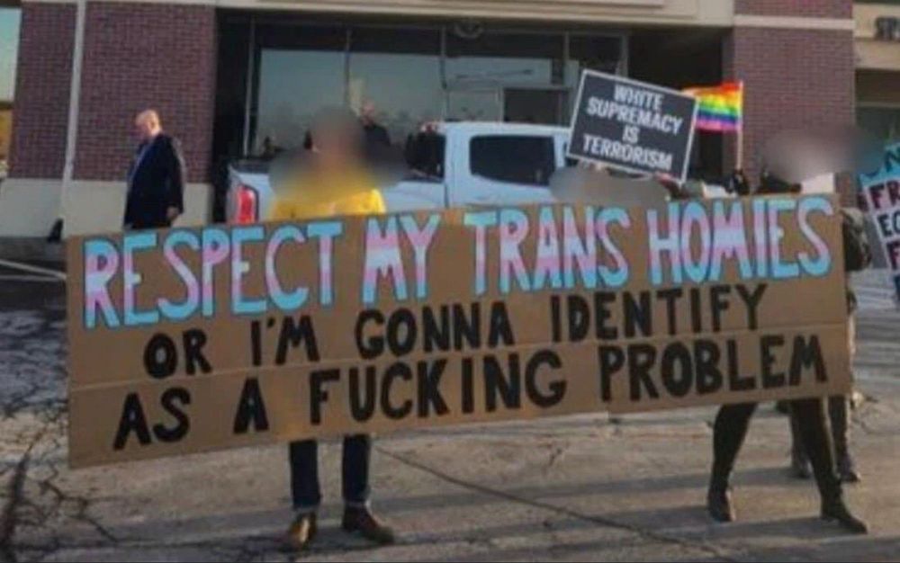 Large banner held by two people. Text of banner is "RESPECT MY TRANS HOMIES OR I'M GONNA IDENTIFY AS A FUCKING PROBLEM". "Respect my trans homies" is painted in the colors of the trans flag; the rest of the text is black. The banner is painted on brown cardboard.