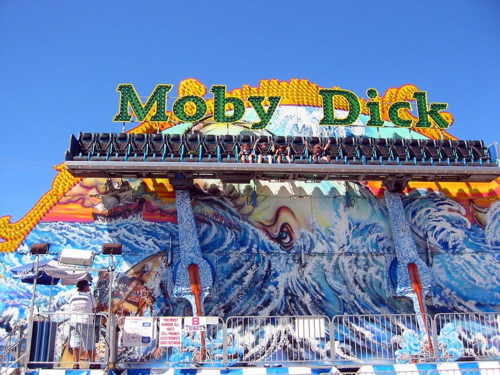 A fair ride that hoists riders strapped into seats up into the air, quite high but not upside down. It’s whale themed with airbrushed white whale art and says MOBY DICK in big brightly lit letters.