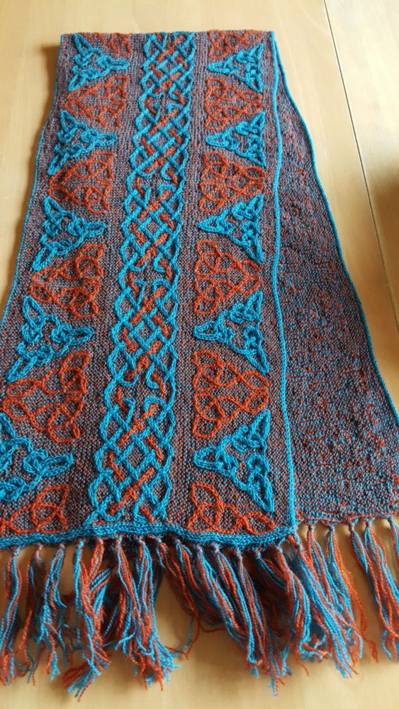 Bleu and red/orange knitted shawl featuring keltic knots on a wooden floor (Lindisfarne)