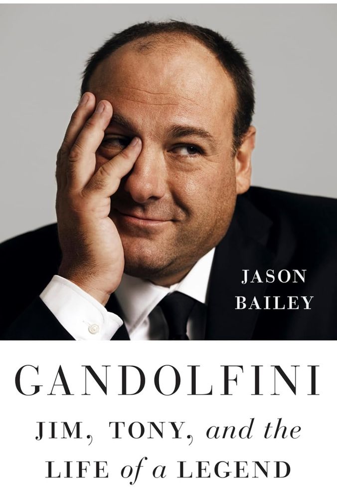 The cover of Gandolfini: Jim, Tony, and the life of the legend, by Jason Bailey. Photograph of actor James Gandolfini in a black suit and white shirt and black tie, leaning his right hand against his face, smiling.
