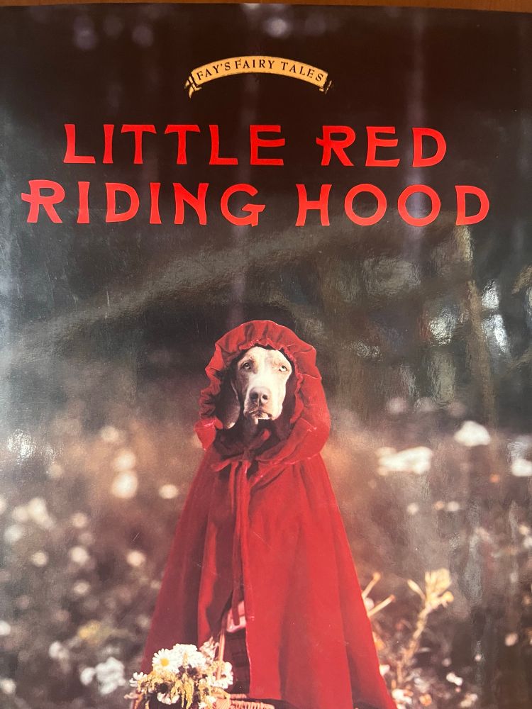 A forlorn looking dog is wearing a red cape under the title LITTLE RED RIDING HOOD