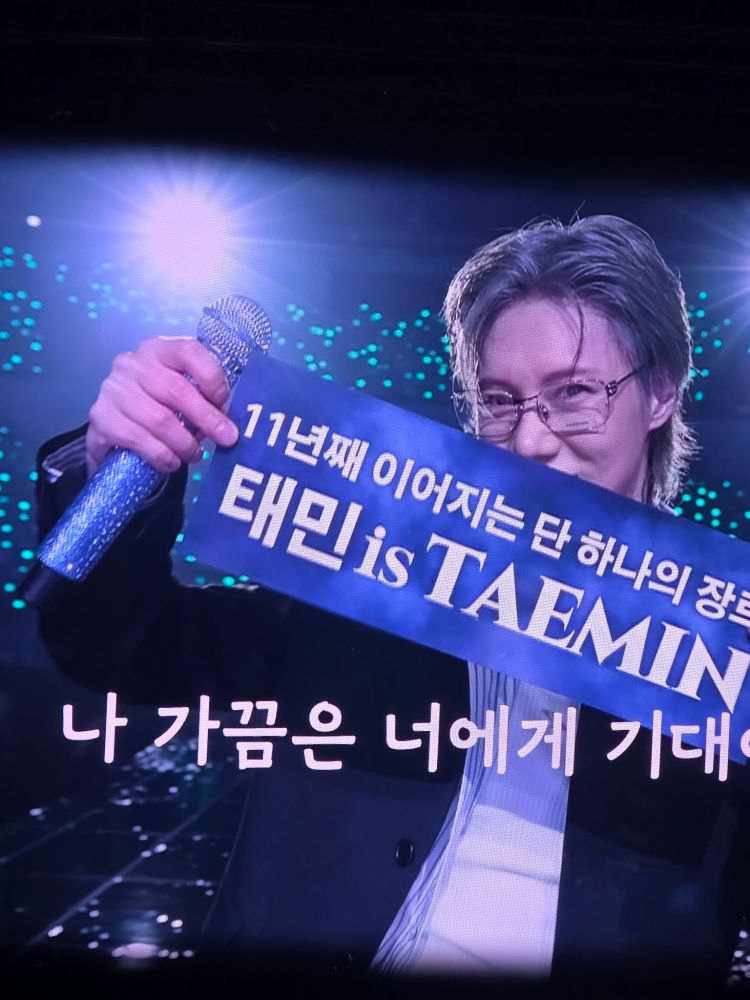 Taemin holding a banner posing for a photo with the audience during his CLASS-MATE fan meeting. He has silver hair combed back and sexy fake glasses. Credit: @letme_go_home on X.