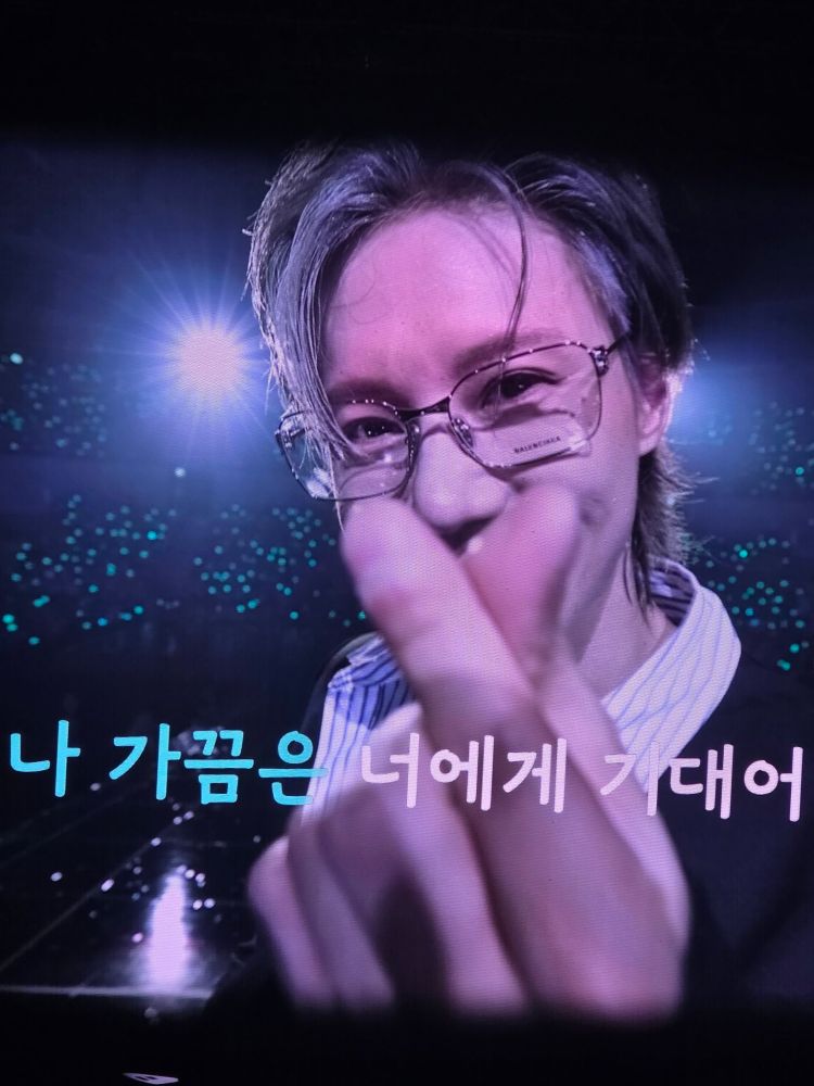Taemin holding up a finger heart. Devastating eye smiles. His glasses have the logo still on the glass. Credit: @letme_go_home on X.