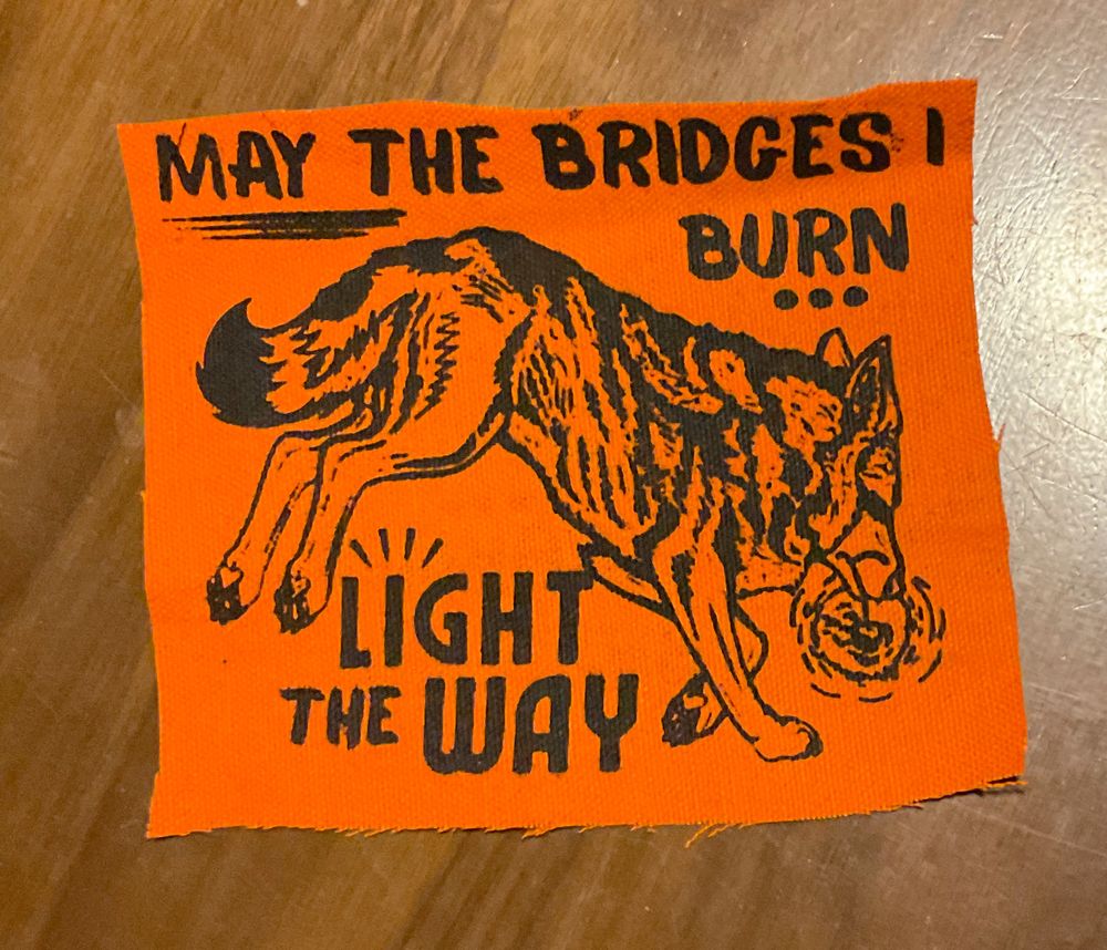 Screen printed patch, black ink on orange fabric, showing art of a coyote running to the right and holding a match in its mouth. It says "May the Bridges I burn light the way"