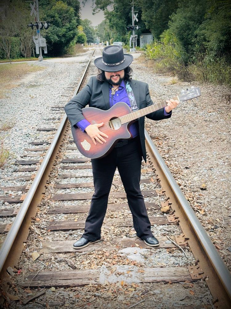 Man standing on the railroad tracks holding a guitar. He is wearing a black suit over a purple western shirt and a black cowboy hat.  His eyes are shadowed by his hat and he is smiling 