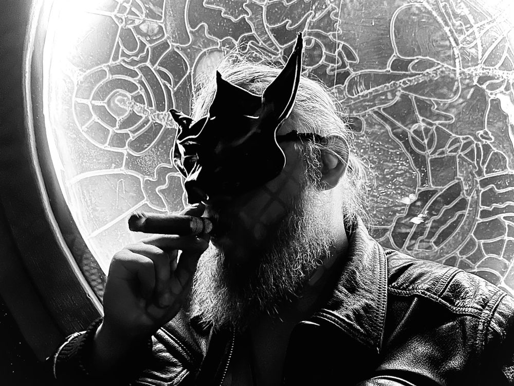 A black and white photo of a gay otter smoking a cigar with a black leather satyr mask on.