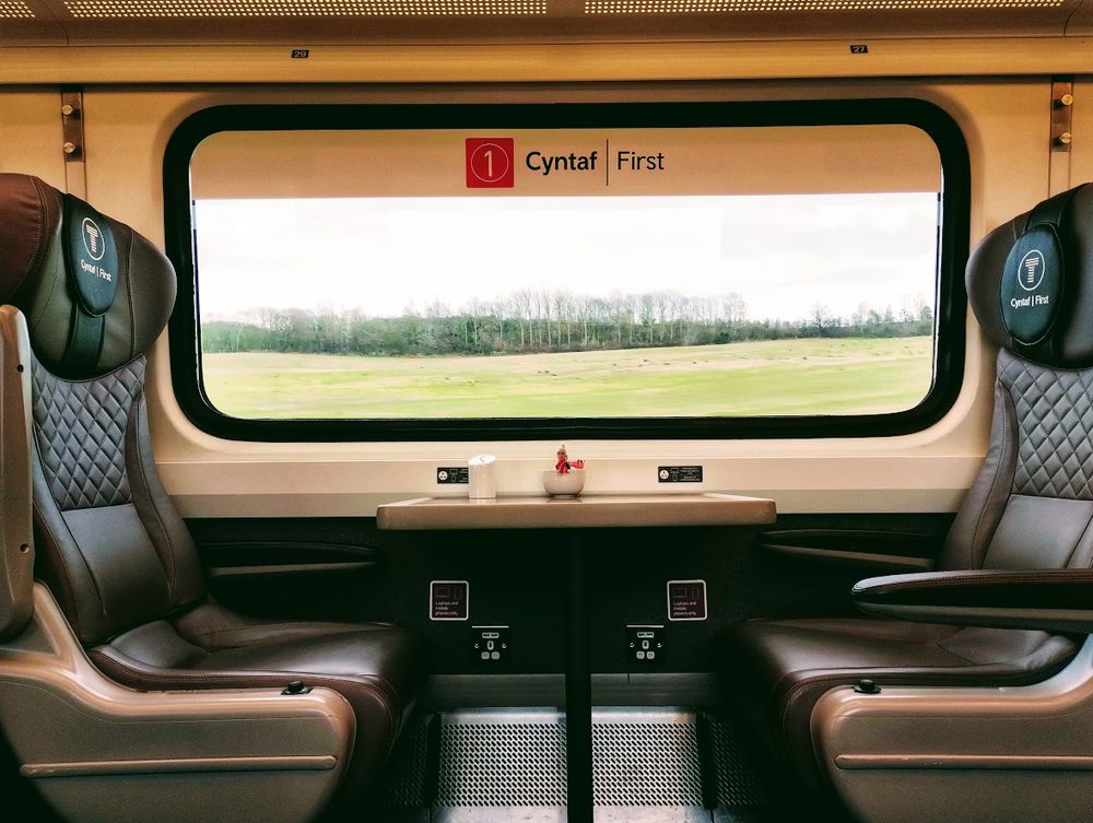 A view of a first class window seat on a Transport for Wales service. Through the window we can see trees on a green horizon. Beneath the window, a table with power sockets. Either side of the table, bit comfy leather seats. A Peruvian finger puppet is enjoying the view.