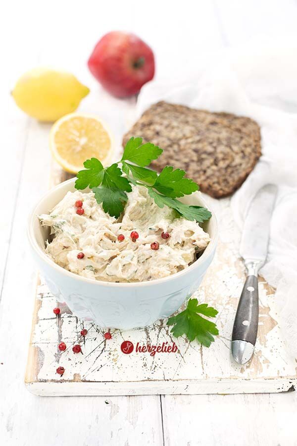 Blaue Schale mit einem Brotaufstrich nach eine Makrelencreme Rezept von herzelieb, garniert mit Petersilie und roten Pfefferbeeren; rechts ein Messer, im Hintergrund zwei Brotscheiben, eine halbe und eine ganze Zitrone sowie ein Apfel.