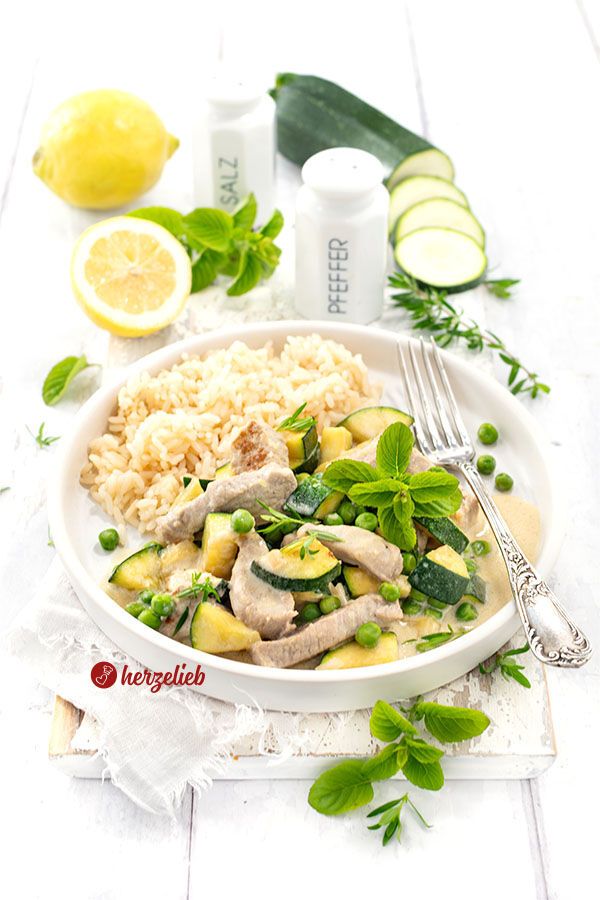 Ein Teller mit Puten-Zucchini-Topf in cremiger Käsesoße, garniert mit frischer Minze und grünen Erbsen, dazu eine Portion Reis. Im Hintergrund liegen frische Zitronen, Zucchinischeiben sowie Salz- und Pfefferstreuer auf einem weißen Holztisch. Ein silberner Tellergabel liegt rechts auf dem Teller.