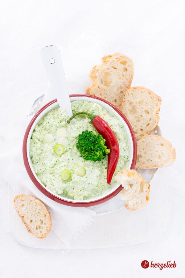 Zartgrüner Brokkoli-Dip in einer weißen Schüssel mit rotem Rand, dekoriert mit einem Brokkolistück und einer Chili. Einweißer Löffel steckt in der Broccomole, dem Frischkäsedip. Rund um die Schale liegen Baguette-Stücke. Die Aufnahme ist von oben gemacht.