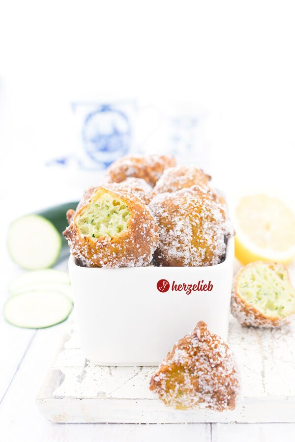 Eine weiße Schale mit goldbraun gebackenen Zucchiniberlinern nach einem Zucchini Berliner Rezept  von herzelieb, die mit Zucker bestreut sind. Ein Kuchenbällchen ist angebissen, grüne Zucchiniraspel im Inneren sind zu sehen. Im Hintergrund frische Zucchinischeiben und eine halbe Zitrone. Links hinten sieht man einen angeschnittene Zucchini und im Hintergrund eine Kaffeetasse.