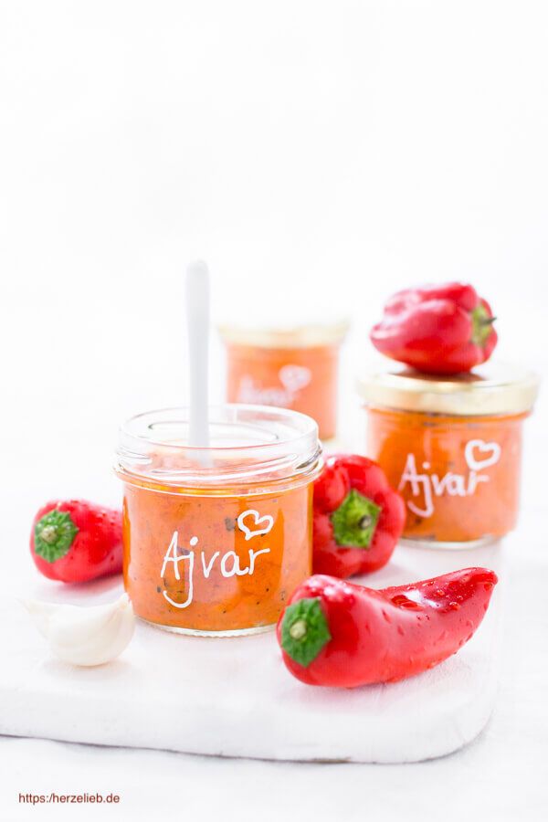 Zwei Gläser mit Ajvar stehen auf einer weißen Fläche, eines davon offen mit einem weißen Löffel darin, umgeben von roten Paprikaschoten und einer Knoblauchzehe; beide Gläser sind mit "Ajvar" und einem Herz handbeschriftet.