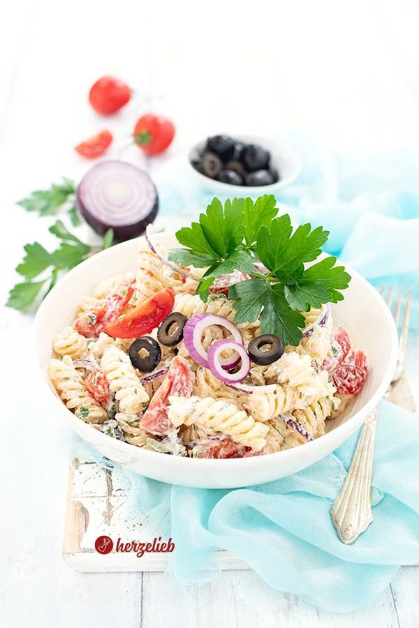Frisch angerichteter Pastasalat nach einem Tzatziki Nudelsalat Rezept von herzelieb in einer weißen Schüssel, garniert mit schwarzen Olivenringen, roten Zwiebelringen, Cherrytomaten und Petersilienblättern, auf einem hellen Holzbrett mit hellblauem Stoff dekoriert. Neben der Schüssel liegt eine Gabel. Im Hintergrund rote Zwiebel, schwarze Olive, Tomaten und Petersilie.