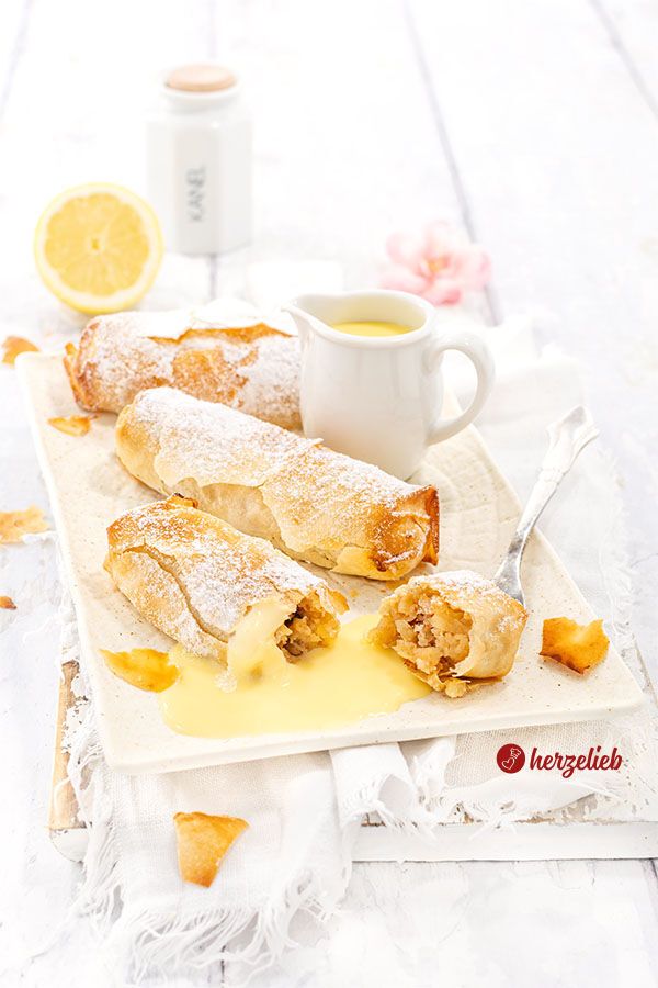 Drei kleine Apfelstrudel, bestäubt mit Puderzucker, liegen auf einem weißen rechteckigen Teller. Einer der Strudel ist angeschnitten, sodass die Apfelfüllung sichtbar wird, die von Vanillesoße umgeben ist. Im Hintergrund steht ein kleines Kännchen mit Vanillesoße und eine halbe Zitrone.