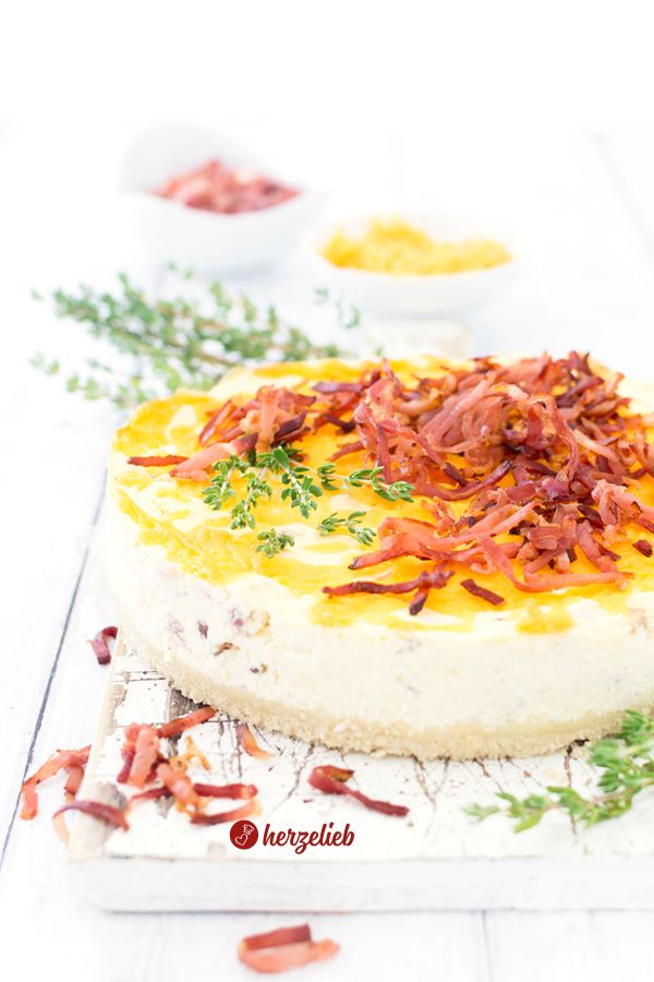 Nahaufnahme eines gebackenen Käse-Schinken-Käsekuchens auf einem Brett, der Bröselboden ist deutlich zu erkennen, oben mit knusprigem Schinken und frischem Thymian dekoriert, umgeben von verstreuten Schinkenstücken auf hellem Untergrund.