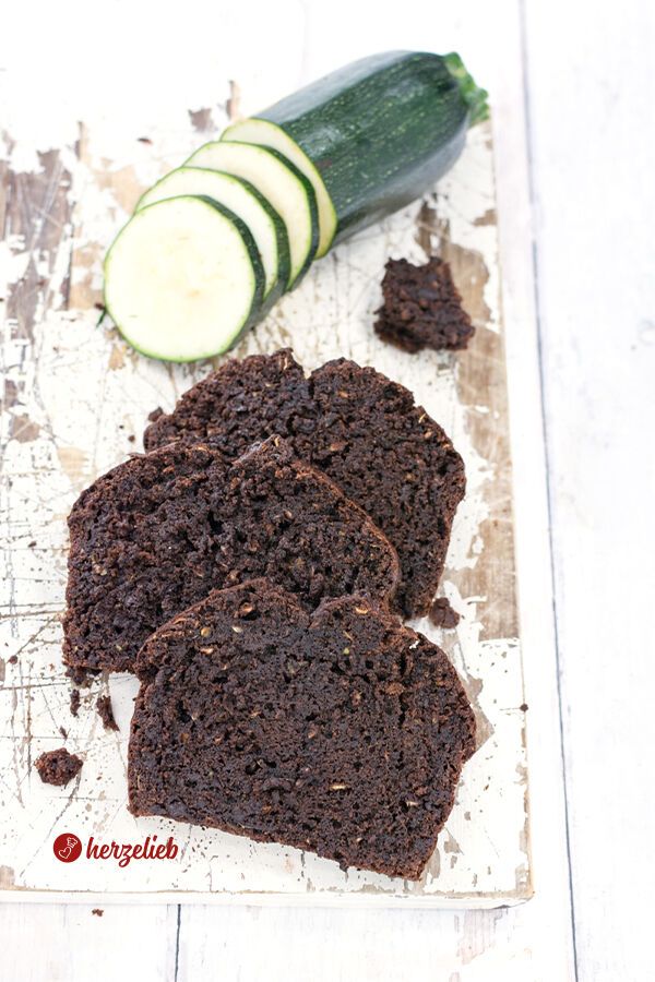 Drei Scheiben von einem dunklen, saftigen Schokoladen-Zucchinikuchen liegen auf einem rustikalen Holzbrett, daneben liegen einige Scheiben frische Zucchini.