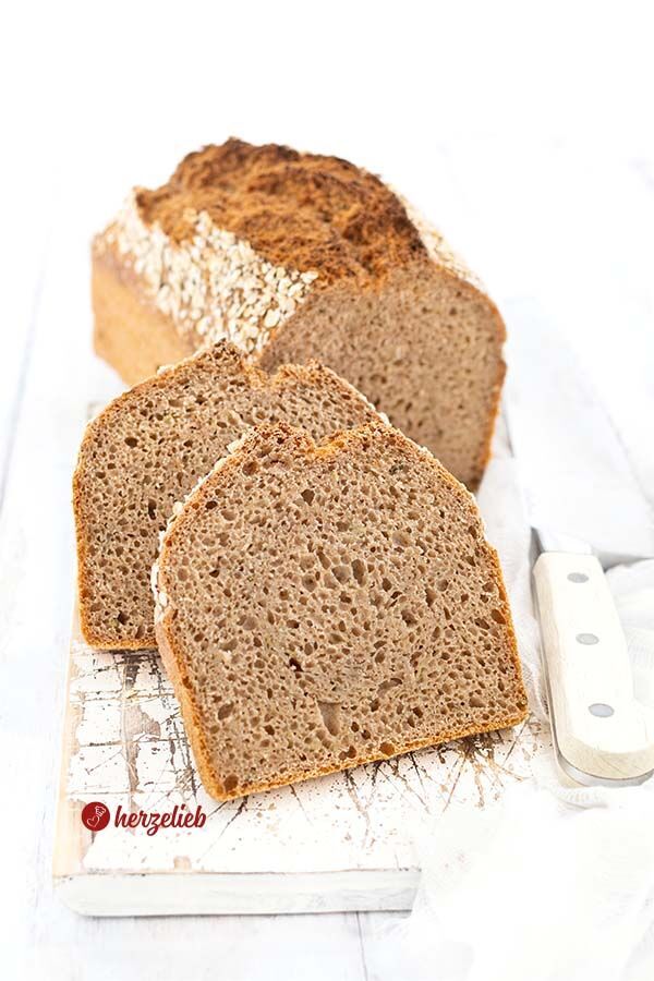 Zwei Scheiben dunkles Zucchinibrot nach einem Vollkorn-Zucchinibrot Rezept von herzelieb sind vor dem Brotlaib auf einem weißen Brett arrangiert, die Kruste von dem Vollkornbrot ist braun und mit Haferflocken bedeckt, rechts daneben liegt ein Brotmesser mit hellem Griff vor hellem Hintergrund.