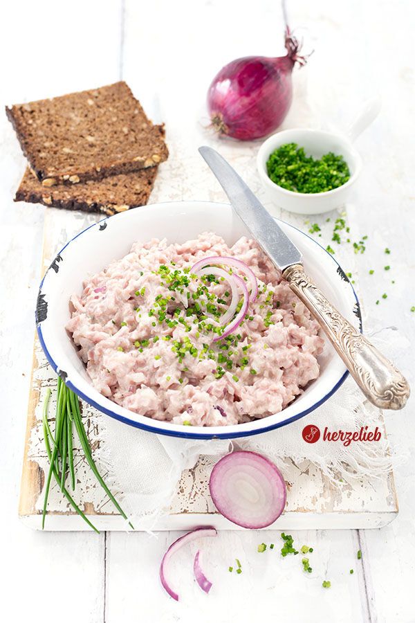 Eine Schüssel mit cremigem Fuglekvidder, einem dänischen Skinkesalat, garniert mit frisch geschnittenem Schnittlauch und roten Zwiebelringen, steht auf einem weißen Holzbrett vor zwei Scheiben Vollkornbrot, einer roten Zwiebel und einem Schälchen Schnittlauchröllchen.