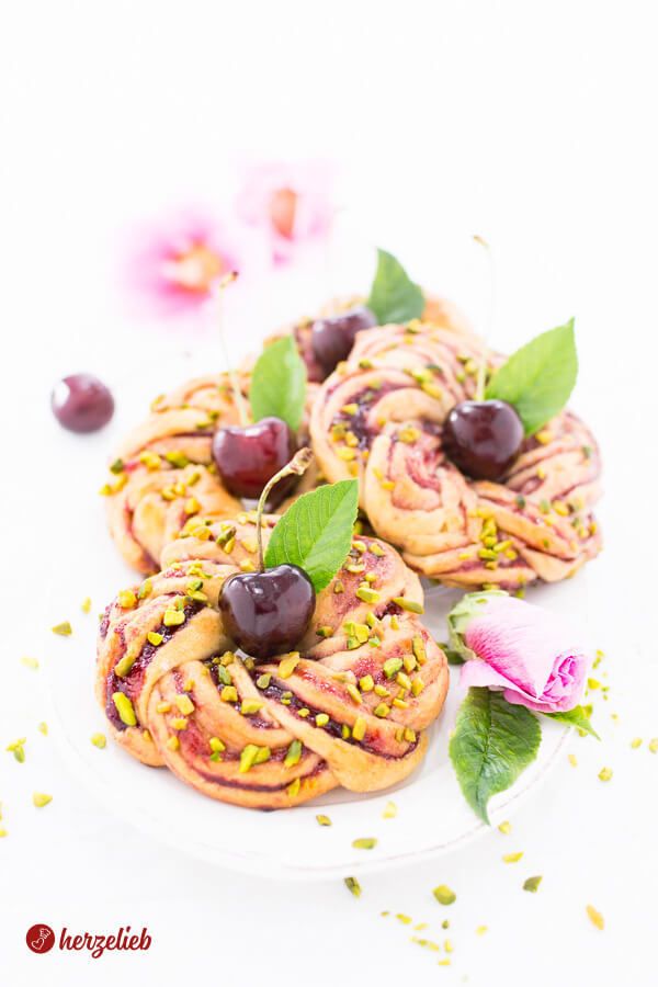 Vier kunstvoll gedrehte Kirsch-Gebäckstücke nach einem Kirschkringel Rezept von herzelieb auf einem weißen Teller, umgeben von verstreuten Pistazienstückchen, frischen Kirschen und einer rosa Blüten mit Blättern.