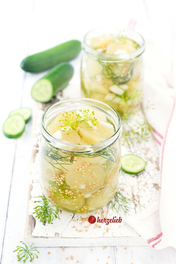 Zwei Gläser mit eingelegten Gurken nach einem Senfgurken Rezept in einer klaren Lake, Zwiebelwürfelchen, Dill und Senfkörnern, auf einem rustikalen weißen Untergrund. Im Vordergrund Dillblüten. Im Hintergrund zwei frische Gurken, eine davon aufgeschnitten. Rechts ein weißes Geschirrtuch mit roten Streifen.
