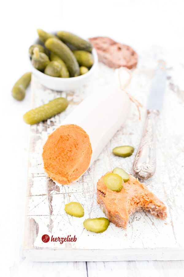 Ein Stück vegetarischer Teewurst-Aufstrich in Wurstform, von dem ein Ende angeschnitten ist. Daneben befindet sich eine Scheibe Brot, die mit dem Aufstrich bestrichen und mit kleinen Gurkenscheiben belegt ist. Einige Gurkenscheiben liegen lose auf der Holzfläche. Im Hintergrund steht eine weiße Schale mit weiteren Cornichons. 