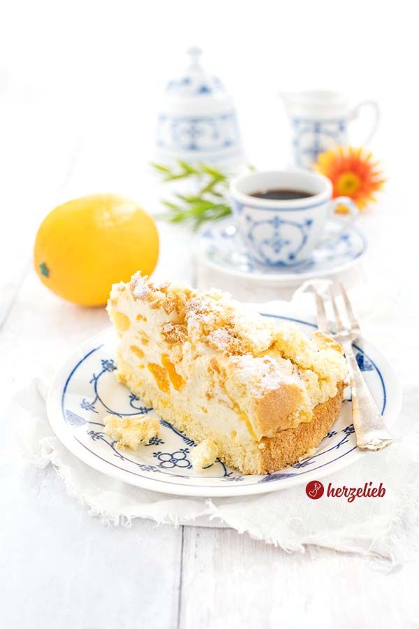 Ein Stück Fruchttorte auf einem blauweißen Kuchenteller sieht man auf diesem Foto zum Mandarinentorte Rezept.Auf dem Tellerrand liegt eine Kuchengabel. Hinter dem Teller links ein Orange, rechts eine Tasse Kaffee. Dahinter ein Zuckertopf und ein Milchkännchen und eine orangefarbene Blüte