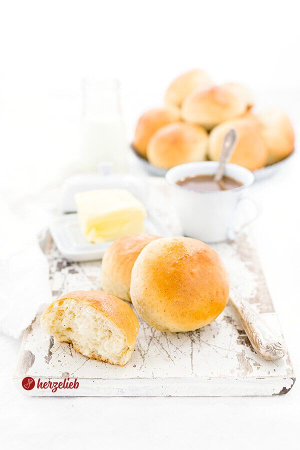 Im Vordergrund liegt ein angebissenes Brötchen. Dahinter sind zwei übereinanderliegende Milchbrötchen und seitlich ein Messer zu sehen. Im Hintergrund stehen eine Tasse Kaffee, eine geöffnete Butterglocke mit Butter sowie ein Korb voller goldbraun gebackener Milchbrötchen.