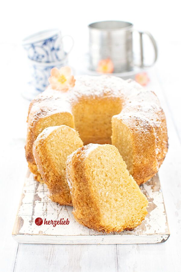 Frisch gebackener Eierlikör-Gugelhupf nach einem Eierlikörkuchen Rezept von herzelieb, angeschnitten und auf einem hellen Untergrund präsentiert. Zwei Kuchenscheiben liegen leicht versetzt vor dem Kuchen, der mit feinem Puderzucker bestäubt ist.