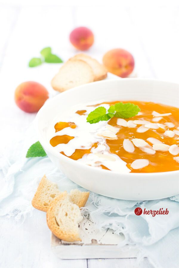 Eine weiße Schüssel mit leuchtend gelbe Aprikosengrütze oder gelbe Grütze, die mit Mandelblättchen bestreut ist. Daneben ist ein Klecks flüssige Sahne oder Milch, dekoriert mit einem frischen Minzblatt. Im Hintergrund liegen drei frische Aprikosen und mehrere Stücke Zwieback auf einem hellen, rustikalen Untergrund.