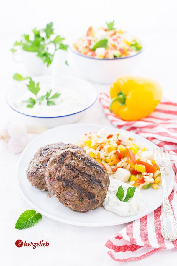 Ein vollständiges Gericht mit zwei gegrillten Frikadellen nach einem Bifteki, Rezept von herzelieb, Kritharaki-Salat und Tzatziki auf einem weißen Teller. Dahinter liegen eine gelbe Paprika, eine Schale mit Tzaztiki und eine mit dem Salat  Knoblauchzehen und frische Kräuter auf einem rot-weiß gestreiften Küchentuch.