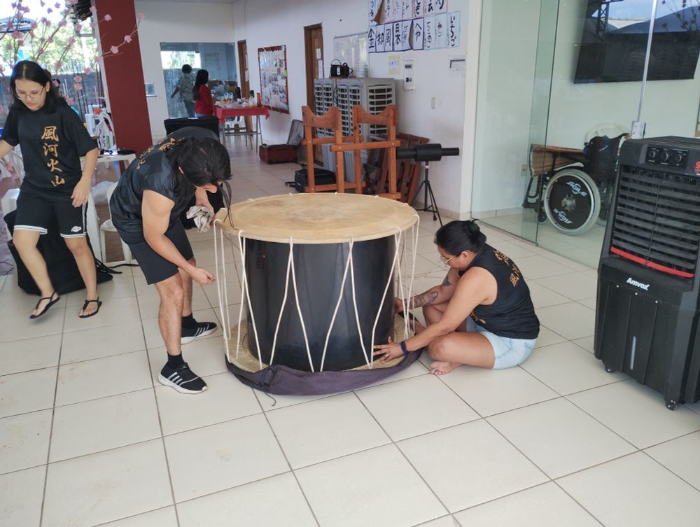 Duas pessoas estão afinando um grande tambor japonês (taiko) em um espaço interno. Uma delas está em pé e a outra sentada, ambas puxando cordas que prendem o couro do tambor. 