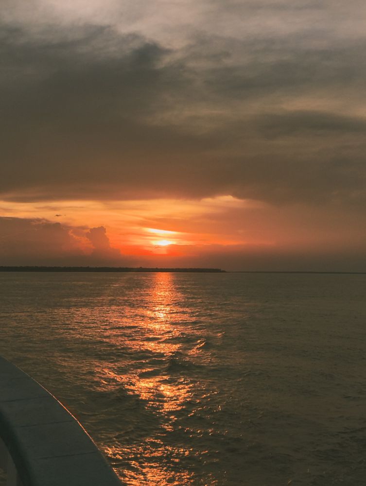 Pôr do sol sobre o rio negro - AM. com o céu alaranjado e nuvens escuras, refletindo tons dourados na superfície da água.
