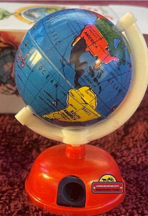A vintage 1960s 3" globe pencil sharpener. A tin painted globe sits at an angle, held by a white plastic semi-circular piece attached to the North and South Poles of the globe. The globe and holder attach on the bottom to a red, domed, pencil sharpener. Note that the tin globe can be detached from the white holder. The globe is 2 pieces, joined at the equator. The 2 pieces can be separated; amazing secrets can be hidden in the globe once you put the 2 pieces back together. The secrets can be held there forever, or rolled across your classroom floor to your best friend.