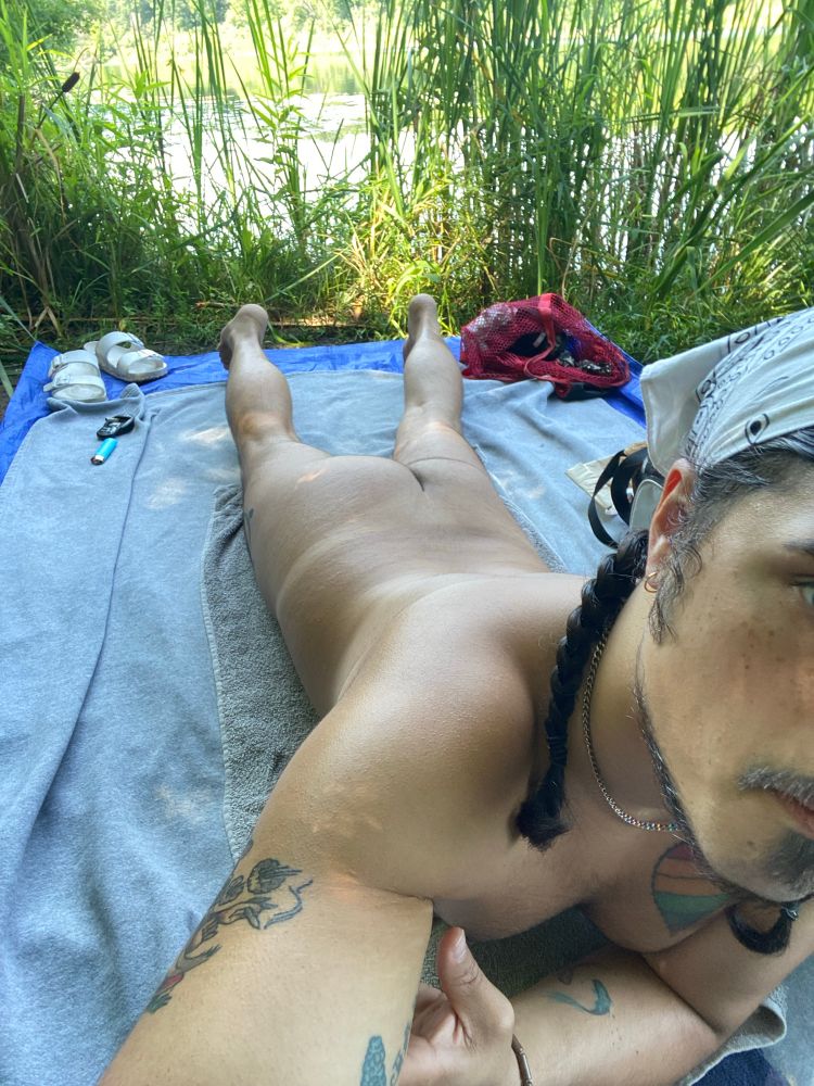 Me laying on my blanket by the water. Some reeds in the back providing some privacy. Laying on my stomach. Shot of a little of my face and then the rest of my body. Ass is up and a little bit of a previous tan line is seen.