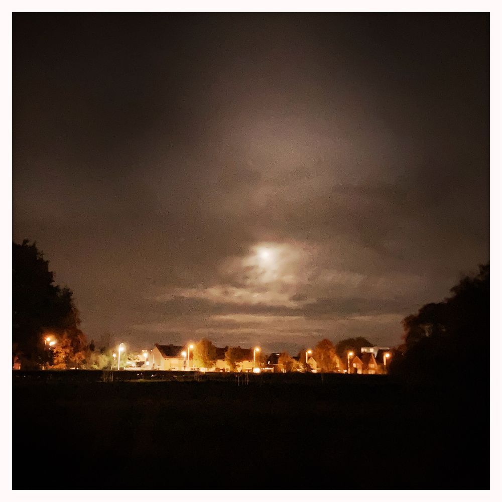 Op de voorgrond een onbelicht veld met aan weerszijden wat bomen, daarachter een rij huizen die geel-oranje belicht zijn en daarboven een bewolkte lucht waar de maan doorheen schijnt. 