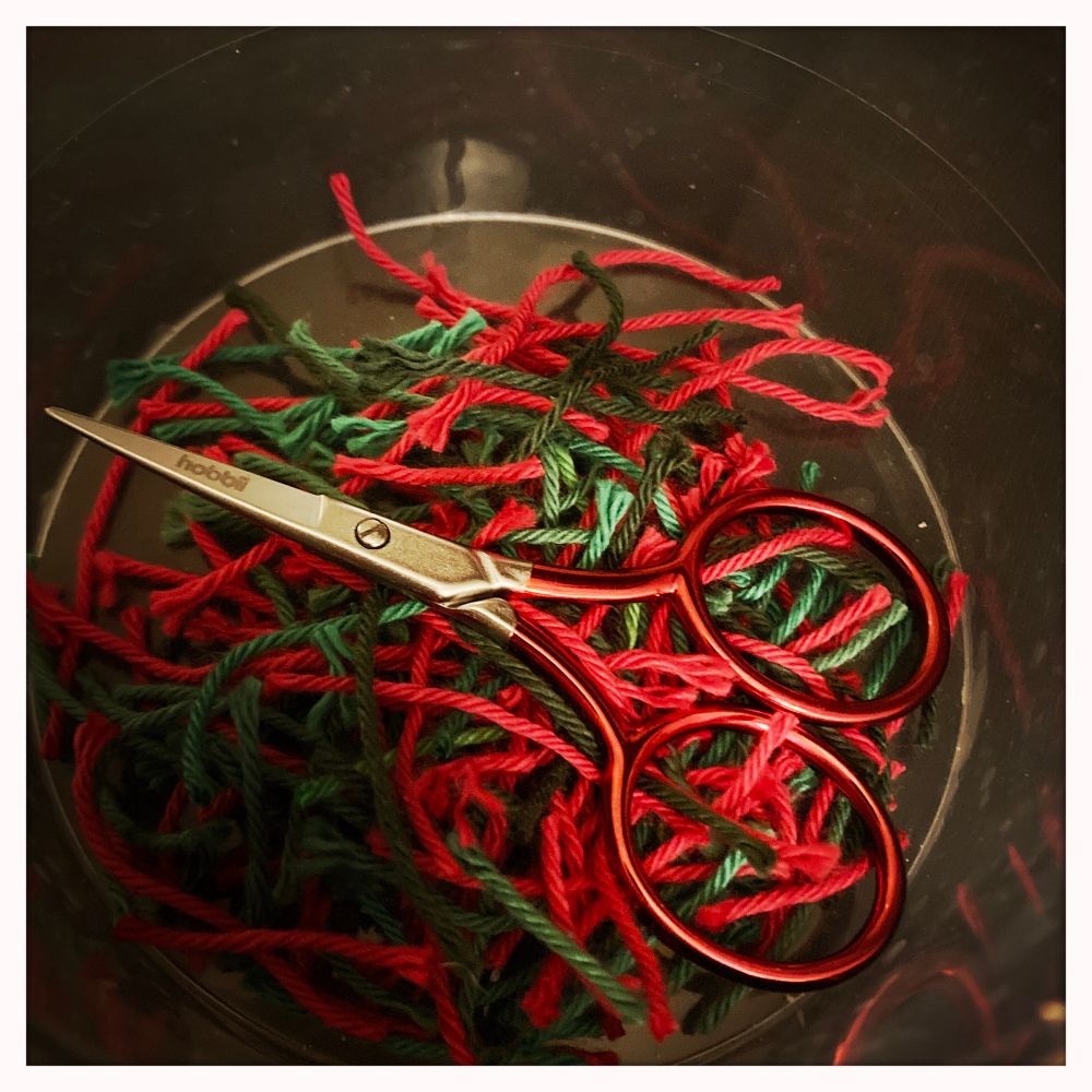 De bodem van een doorzichtig plastic bakje. In het bakje liggen heel veel korte stukjes garen in rood en verschillende tinten groen. Bovenop de draadjes ligt een klein puntig schaartje met metallic rode ogen. 