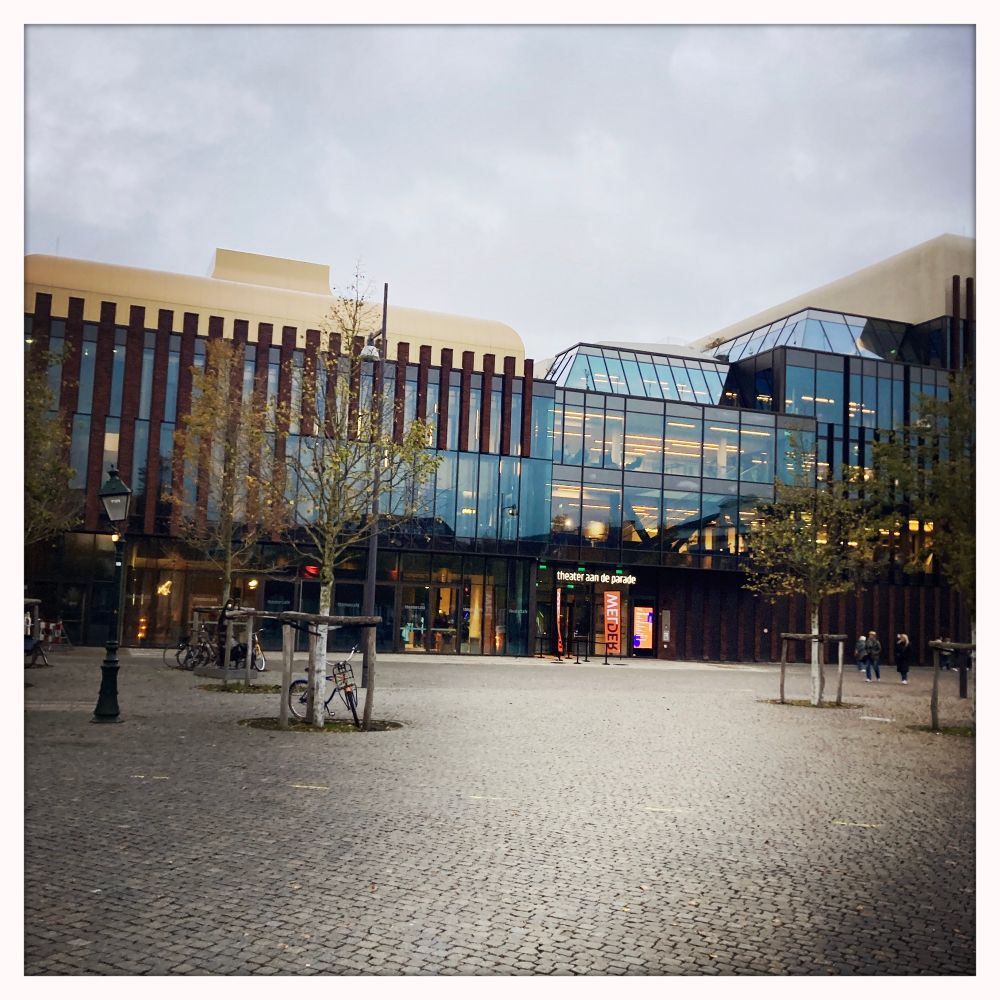 Theater aan de Parade gefotografeerd vanaf de Parade. Op de Parade staan een paar jonge bomen (bij een paar staan fietsen geparkeerd), en een ouderwetse lantaarnpaal. 