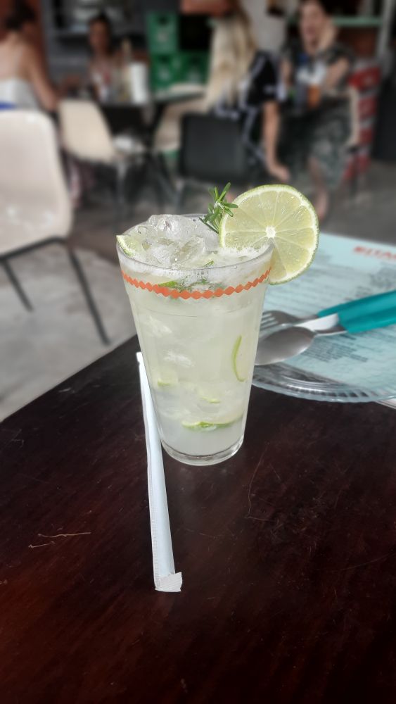 Na imagem aparece um copo grande, cheio de gelo e de líquido. No líquido tem uma mistura de limão, açúcar e cachaça. O copo está decorado com uma rodela de limão. Ao fundo aparece um garfo, uma faca e uma colher em cima de um prato.