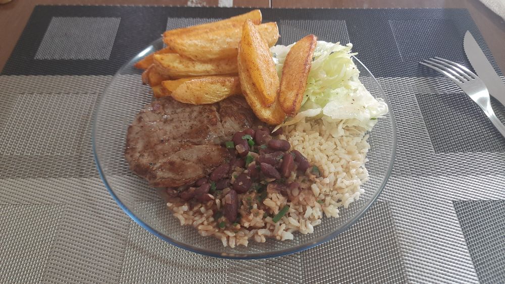 Na imagem aparece um prato de vídeo transparente e dentro dele tem arroz, feijão, bife, salada e batata assada. Ao lado tem um garfo e faca. 