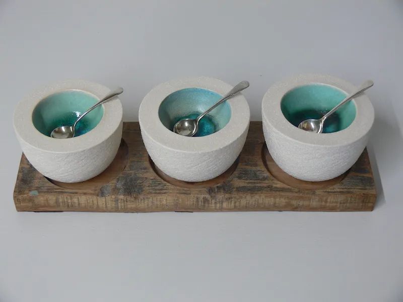A photo of Rhian Winslade's ceramic salt pots. Three resting in a wooden base. They are a light coloured clay, almost white, with a blue glaze inside. They are lined up horizontally with a silver spoon in each.
