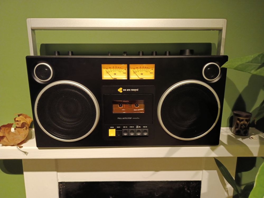 This is a photograph of a beat box, basically. It's a new machine made by the company We Are Rewind. It's mainly black with silver trim. Two large round speakers are on either side of the front with smaller round speakers just above them. There are two VU dials with needles that are lit up in the centre of the top of the front beneath them is the We Are Rewind logo and the actual cassette deck. Various buttons for controlling it are underneath. It is sitting on a mantelpiece, which at the moment is the only place I can find for it. There is a painted green wall behind. The mantelpiece is white. Also on the mantelpiece is a dried leaf and a small ceramic pot, and you can see a few leaves and shadows of leaves on the right-hand side. 