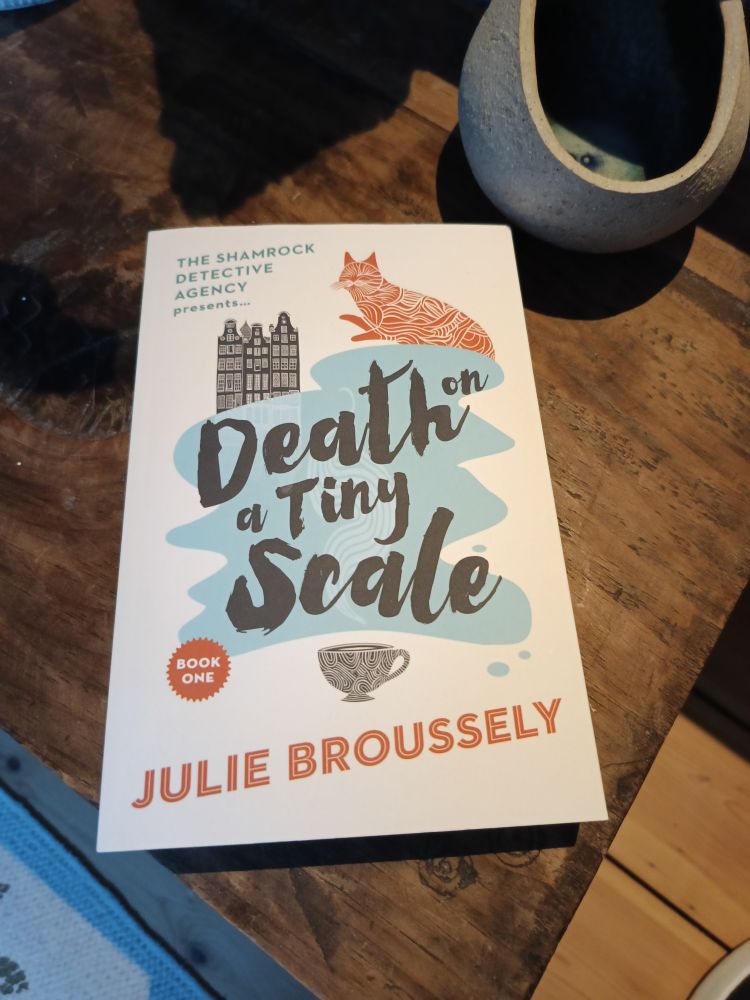 A paperback copy of a novel called Death on a Tiny Scale by Julie Broussely. The title of the book features in the middle of the cover in an illustrated font over a drawing of some water, possibly a river or pond. On the outside of the water is an illustration of a cat, a building, and a cup in the top left-hand corner. There is some text that says "The Shamrock Detective Agency presents", and at the bottom left of the illustration it says "Book One". The book is sitting on the top of a wooden chest, and a ceramic bowl is visible in the top right-hand corner of the photo. 
